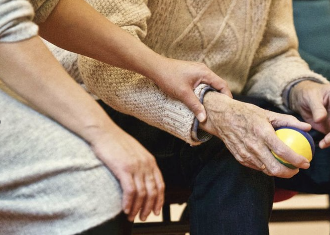 Fotografía de las manos de una persona mayor con una pelota de goma, asistido por las manos de un cuidador